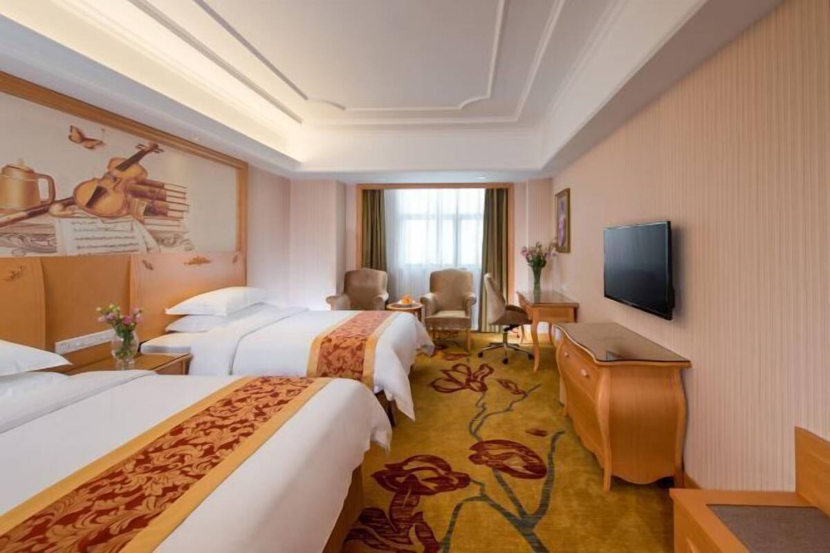 A hotel room with two beds, a TV, a desk, a chair, and a window with curtains.