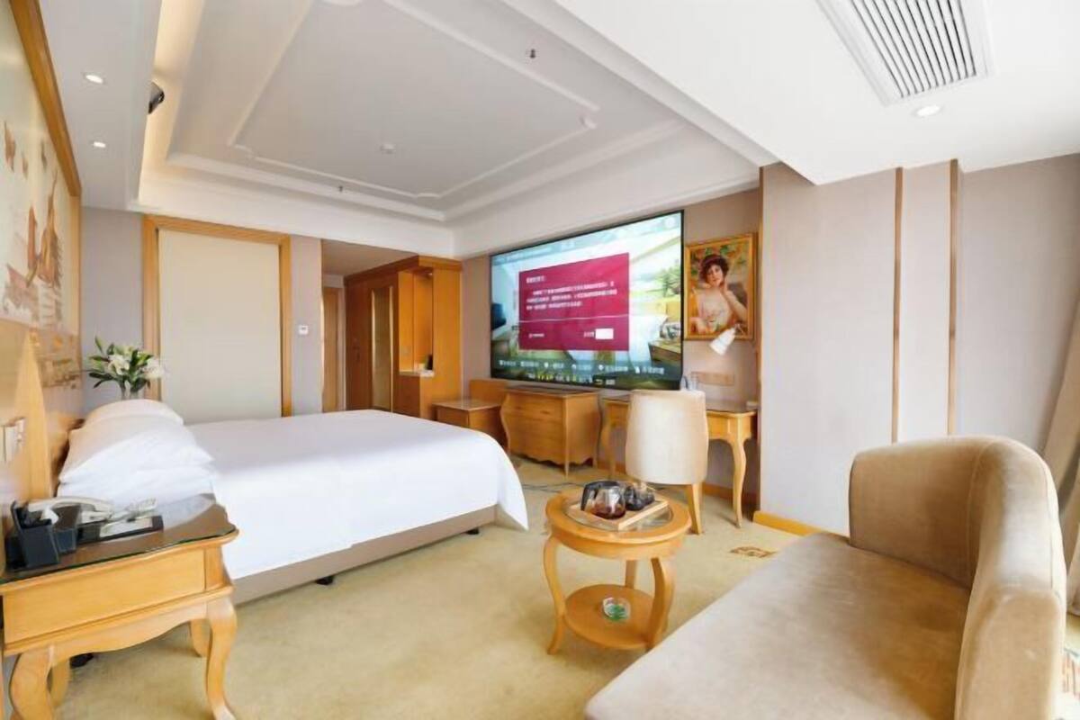 A hotel room with a bed, a desk, a TV, a sofa, and a small table with a vase.