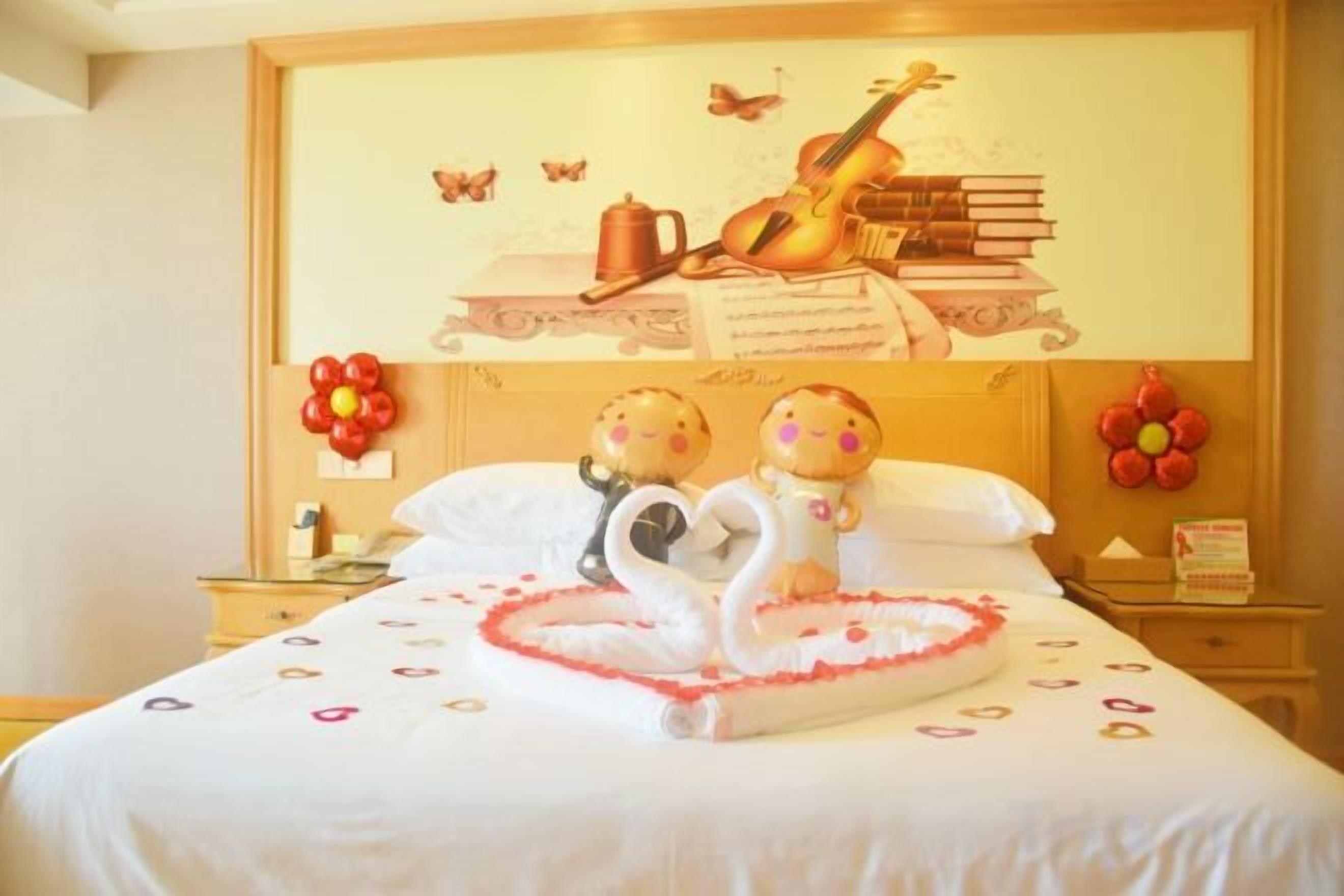 Cama bem arrumada com arranjo de almofadas em formato de coração, parede com instrumento musical e livros, e mesa pequena com um livro e uma decoração de flor vermelha.