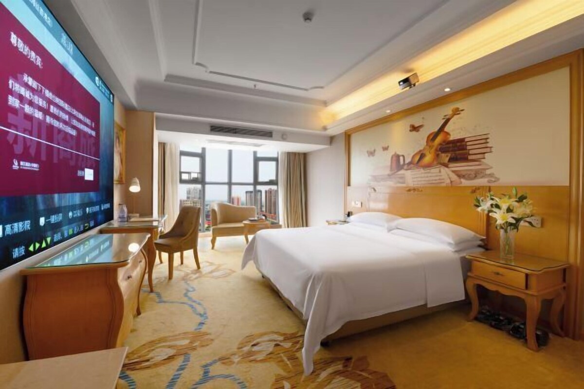 Une chambre d’hôtel avec un grand lit, un bureau doté d’un téléviseur à écran plat, une chaise et une table de chevet agrémentée d’un vase de fleurs.