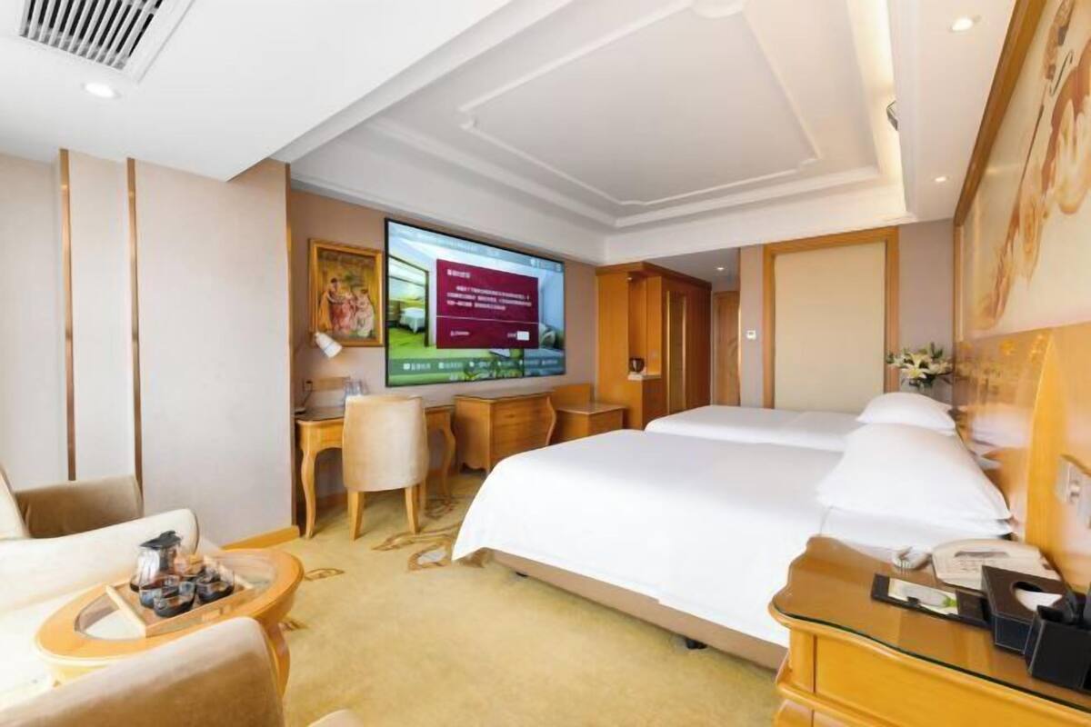 A hotel room with a large bed, a desk with a chair, a television, and a small table with a tray of items.