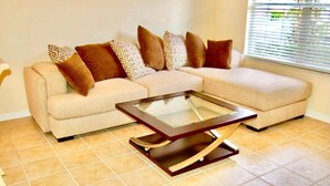 Un salon comprenant un canapé d’angle, une table basse avec un plateau en verre et une fenêtre équipée de stores.