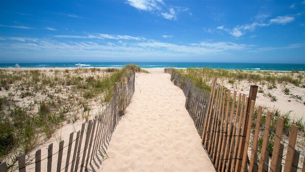 A sandy beach path leading to the ocean, flanked by wooden fences and sparse vegetation.