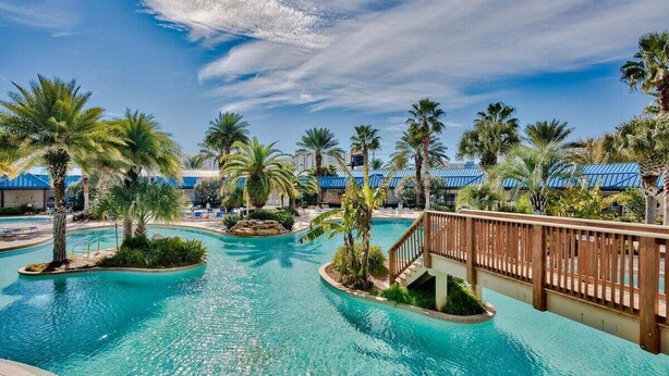 A resort pool area with multiple pools, palm trees, and a wooden bridge. - A Wave Away 1206-Near Beach-Sleeps 6 (Destin)