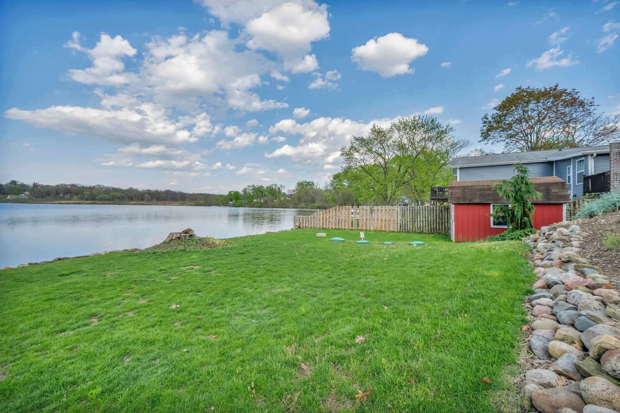 A grassy yard with a wooden fence, a red shed, and a house by a lake.