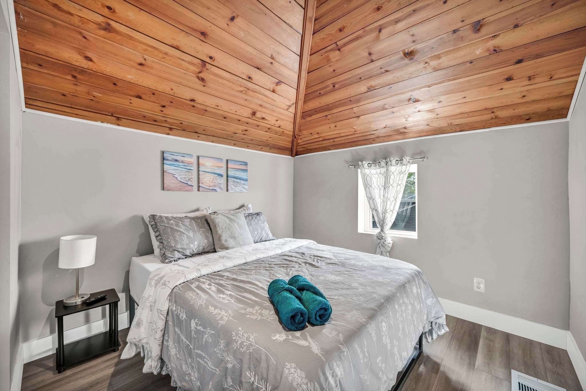 Une chambre avec un plafond en bois, un lit recouvert d’une couvre-lit à motifs floraux gris, deux serviettes enroulées, une table de chevet avec une lampe et une fenêtre avec un rideau.