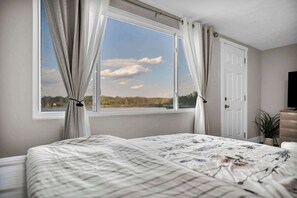 Quarto com cama, janela com cortinas, porta e televisão sobre um penteoulo.