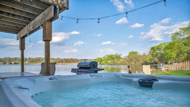 A hot tub with a grill and string lights, overlooking a lake.