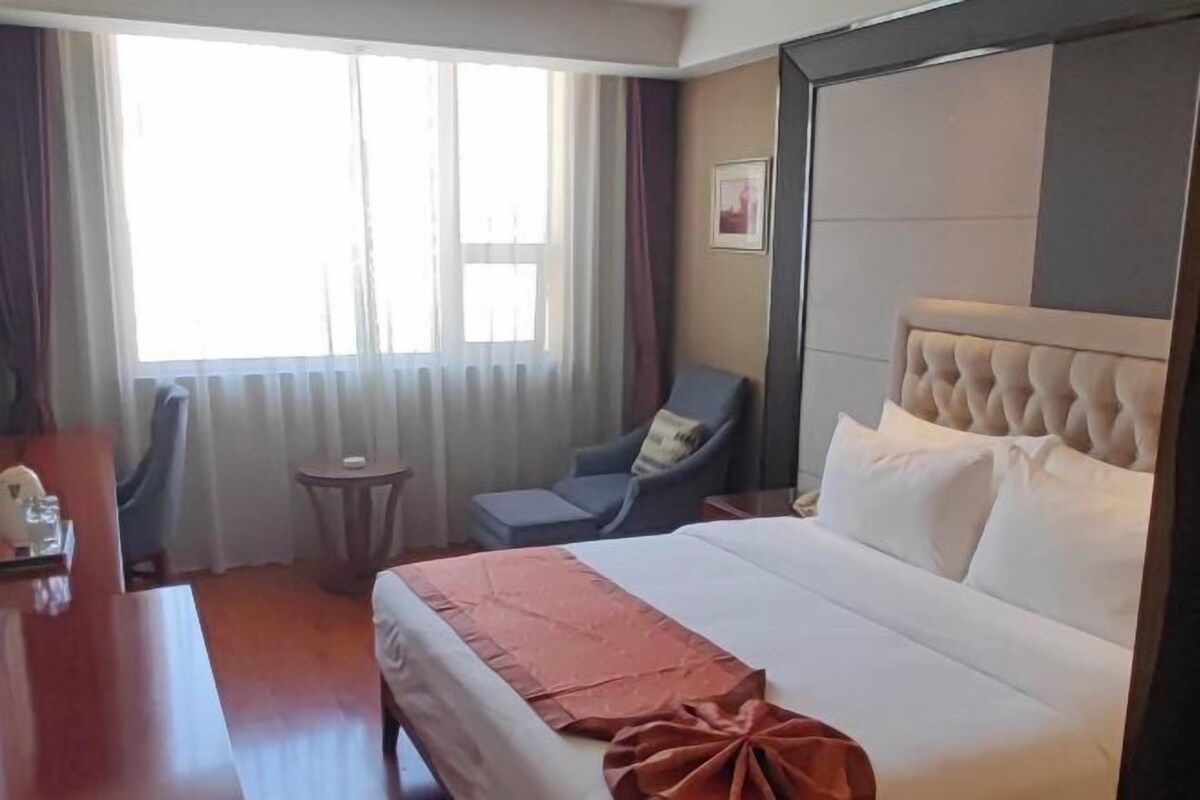 A hotel room with a bed, a chair, a small table, and a window with curtains.