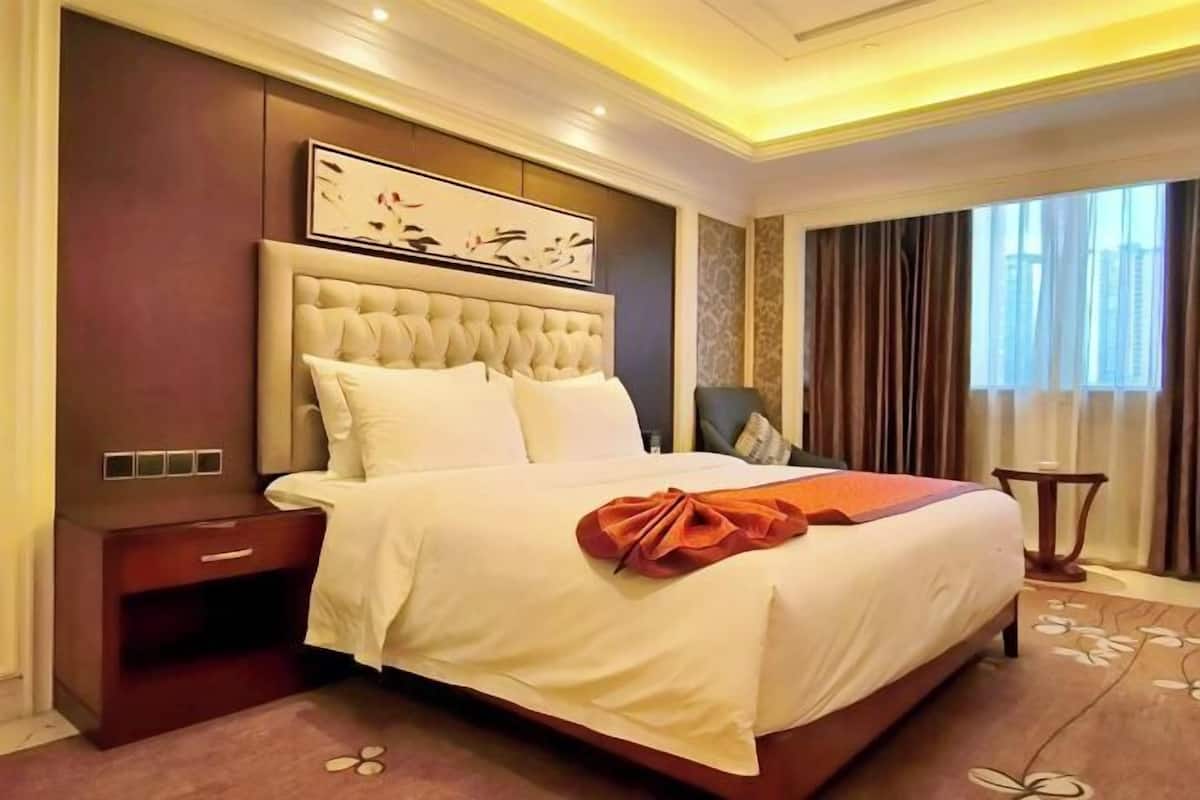 Une chambre d’hôtel avec un grand lit, une table de chevet, une œuvre d’art encadrée au-dessus du lit, une fenêtre avec des rideaux et une moquette à motifs floraux.