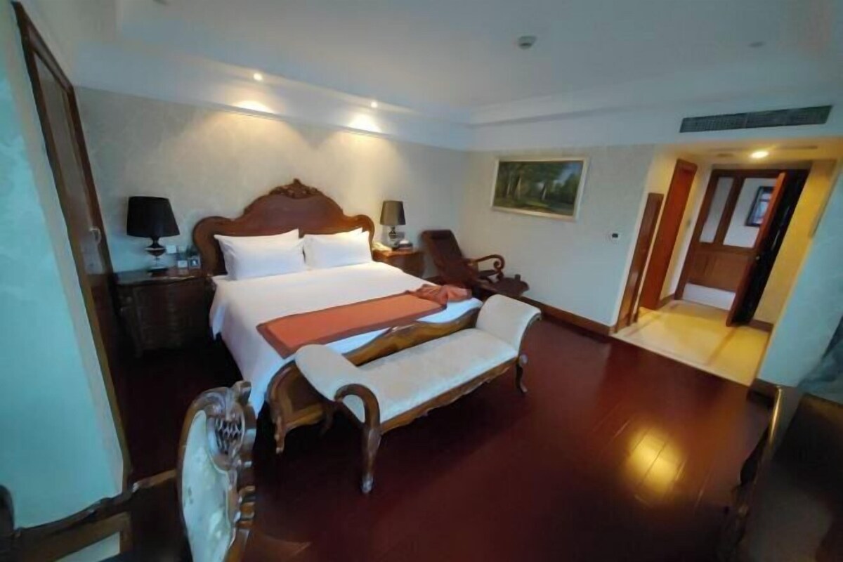 A hotel room with a large bed, a wooden bench, a chair, a nightstand, and a painting on the wall.