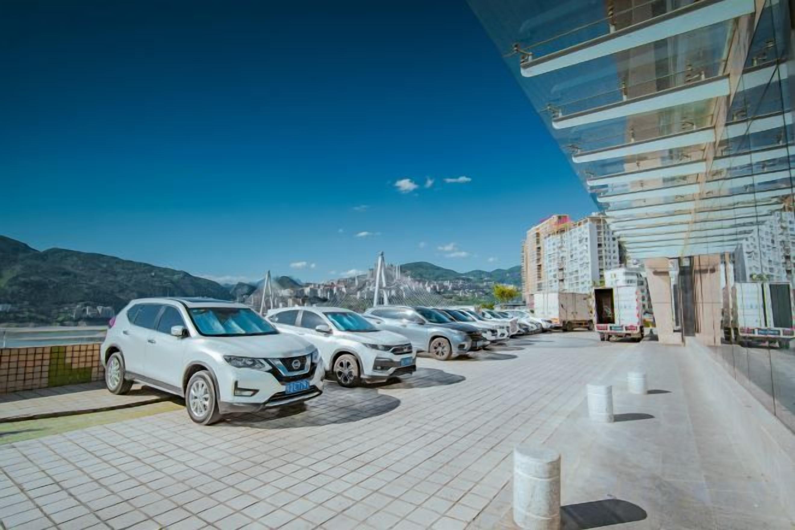 A parking area with multiple cars, a modern building with reflective glass windows, and a mountainous landscape in the background.