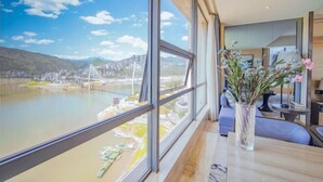 Une chambre moderne avec de grandes fenêtres donnant sur une rivière et un pont, un vase rempli de fleurs et une table soigneusement dressée.