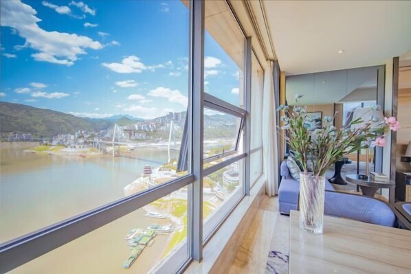 Una habitación moderna con ventanas amplias que dan a un río y un puente, un florero con flores, y una mesa ordenada.