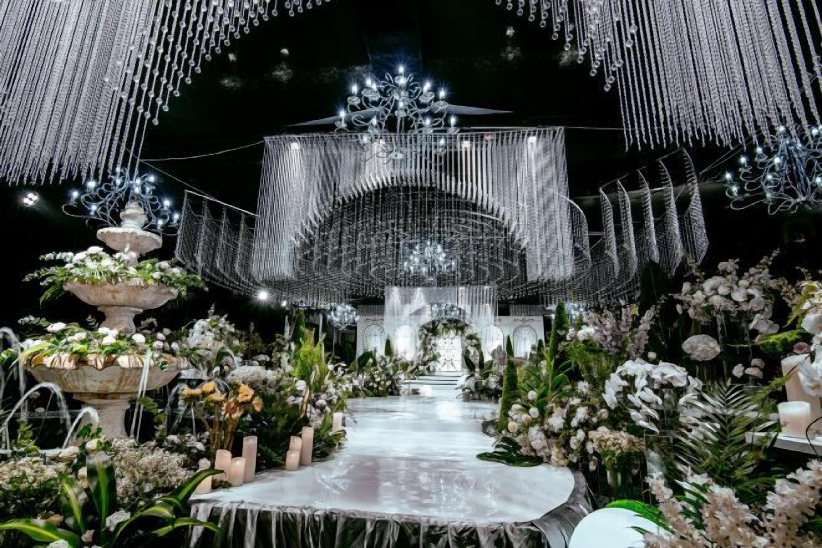 An indoor venue with a central stage, surrounded by floral arrangements and a pathway leading to it.