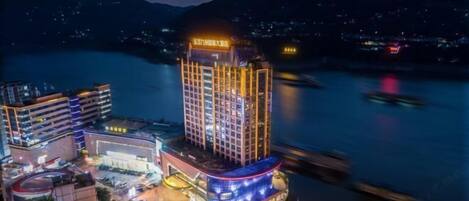 A high-rise hotel with multiple floors, a prominent entrance, and a view of a river and mountains in the background.