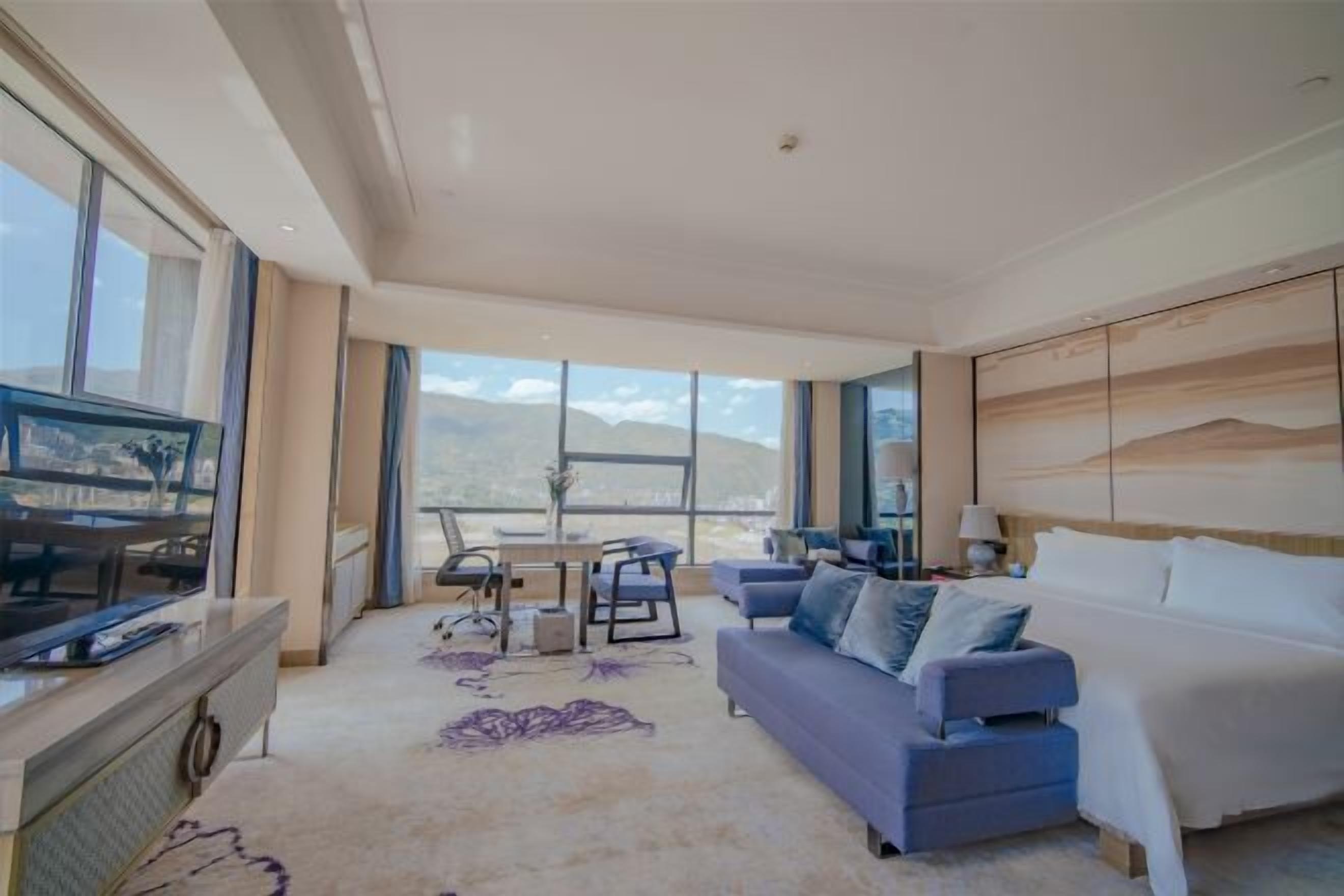 A modern hotel room with a large bed, a sofa, a dining table with chairs, and a TV.