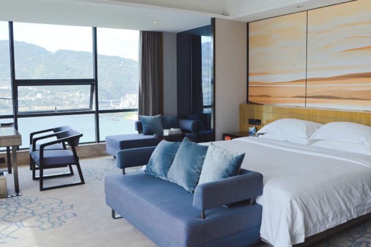 Ein Hotelzimmer mit Bett, Sofa, Schreibtisch und einem großen Fenster mit Blick auf die Stadt.