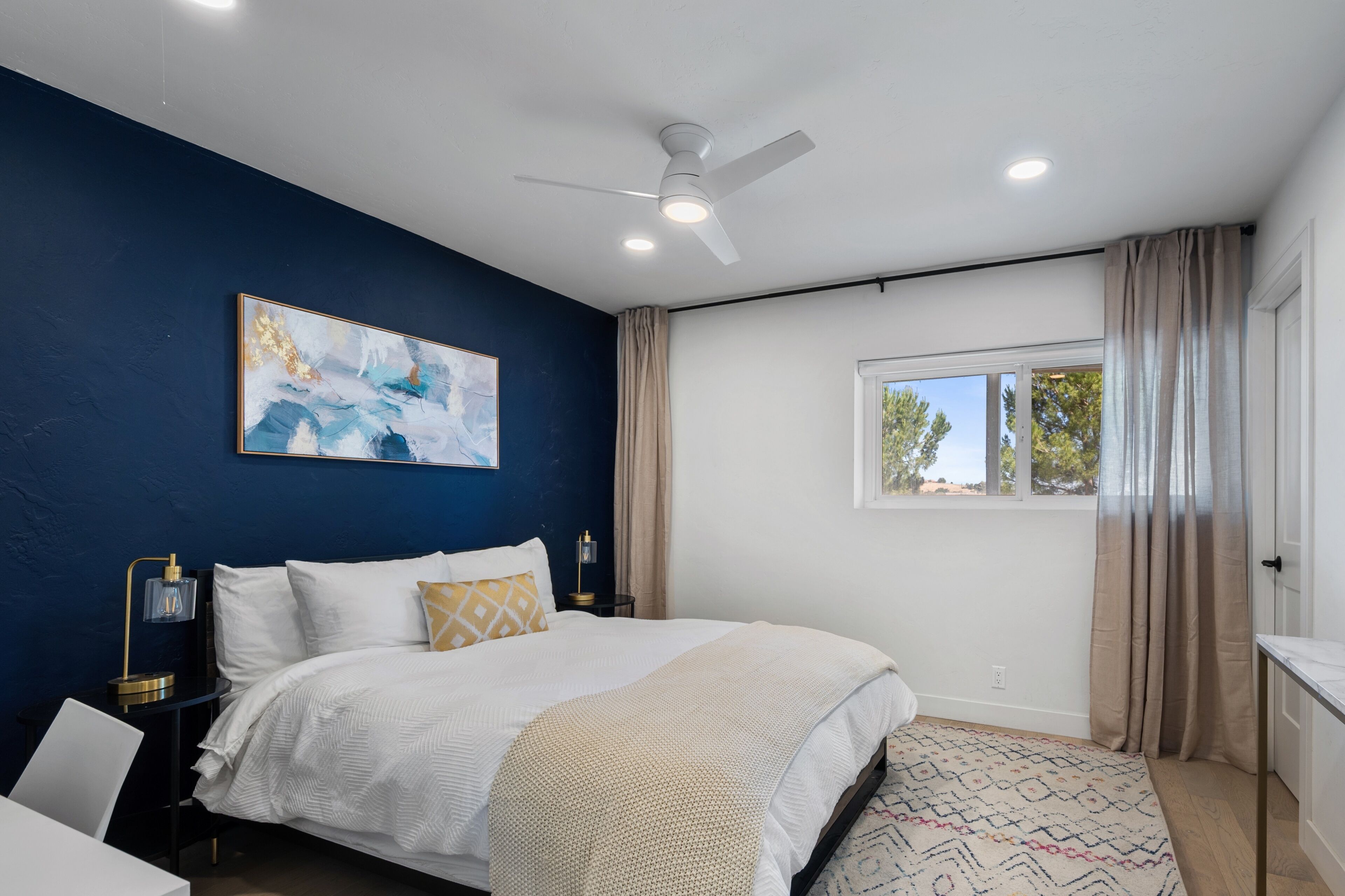 A bedroom with a bed, a ceiling fan, a window with curtains, and a painting on the wall.