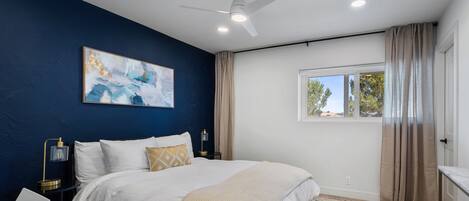 A bedroom with a bed, a ceiling fan, a window with curtains, and a painting on the wall.