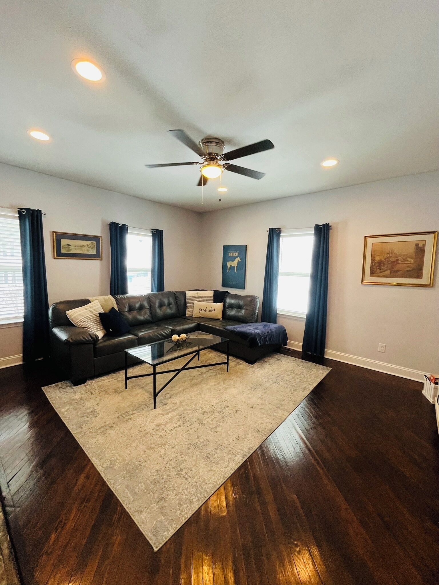 A spacious living room with a dark wood floor, a black leather sofa, a glass coffee table, and a ceiling fan.