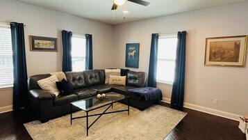 A spacious living room with a dark wood floor, a black leather sofa, a glass coffee table, and a ceiling fan.