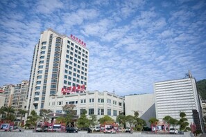 A multi-story hotel with a prominent red sign, a large glass building to the right, and several smaller structures in the foreground. - Lian Shan Longshan Garden Hotel (Lianshan)