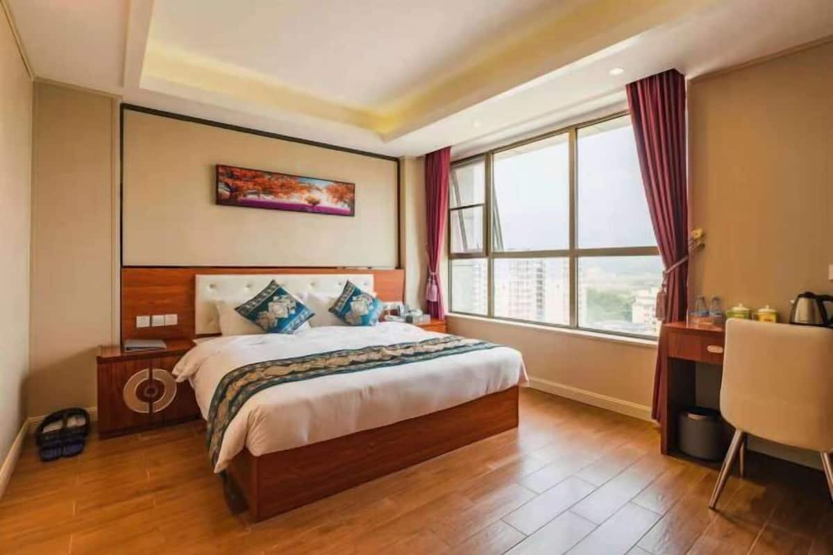 A hotel room with a large bed, a desk, a chair, a window with curtains, and a painting on the wall.