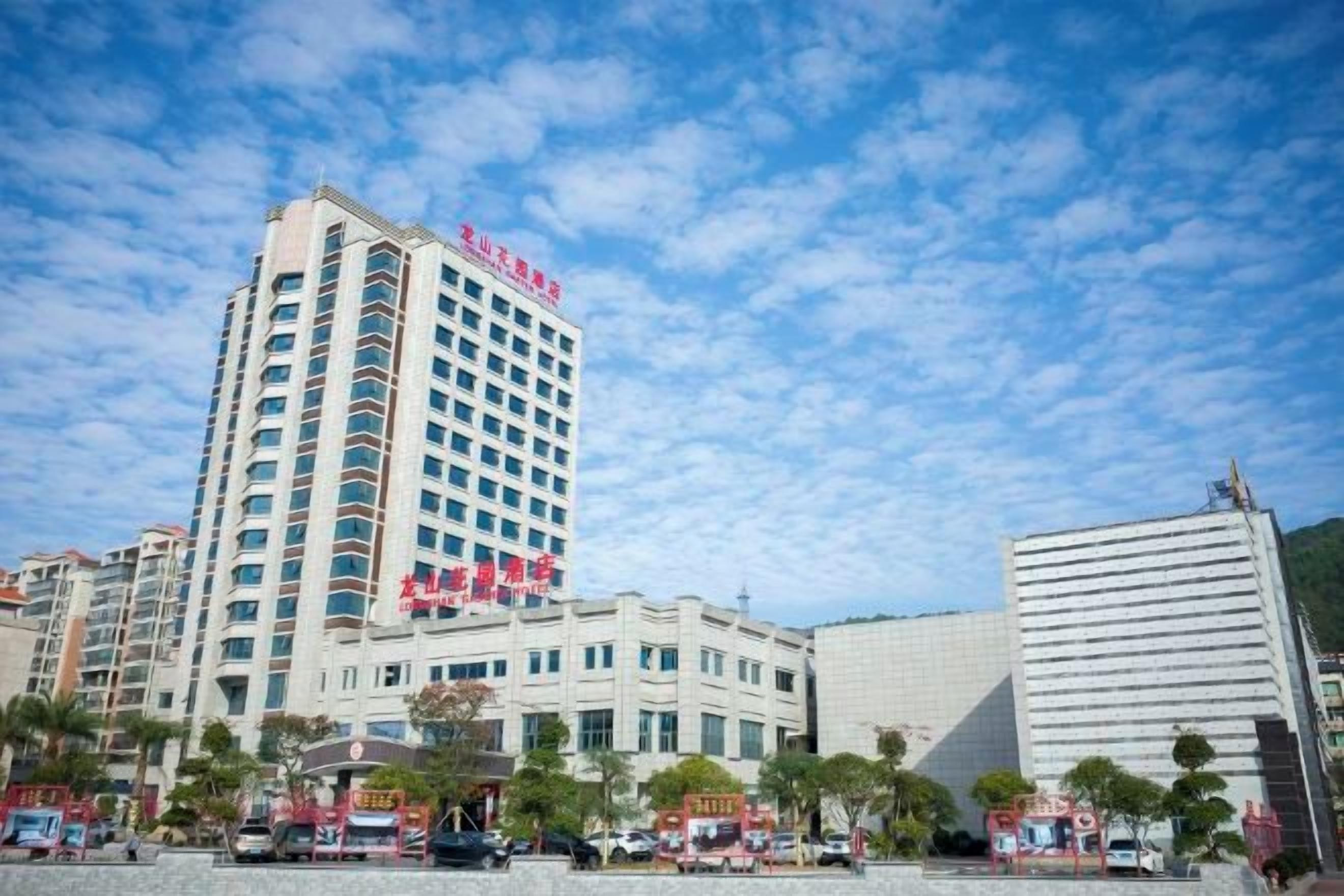 A multi-story hotel with a prominent red sign, a parking lot with several vehicles, and a clear blue sky.