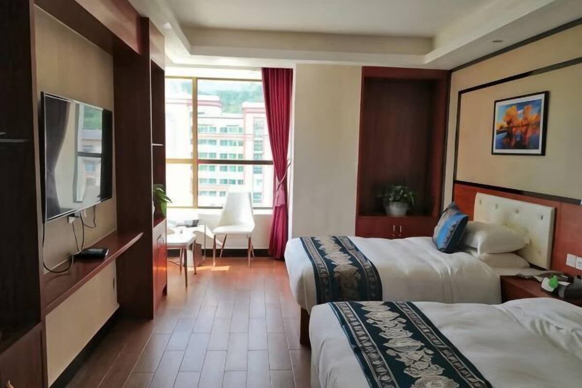 A hotel room with two beds, a TV, a desk, and a window with a view of buildings.