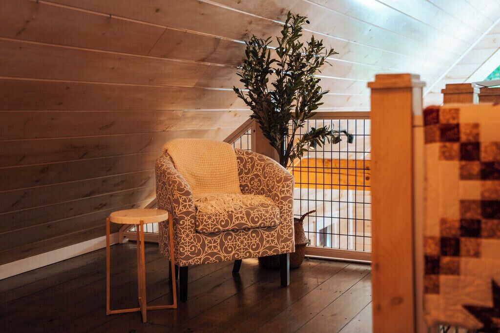 A cozy room with a patterned armchair, a small wooden stool, and a potted plant.