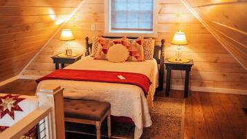 A cozy bedroom with a sloped ceiling, a bed with a red blanket, two bedside lamps, and a small bedside table with a clock.