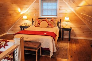 A cozy bedroom with a sloped ceiling, a bed with a red blanket, two bedside lamps, and a small bedside table with a clock.
