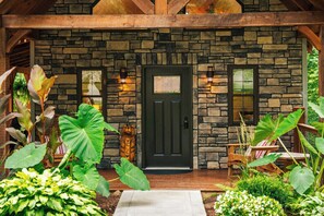 A stone building with a dark door, two windows, and a wooden porch with chairs.