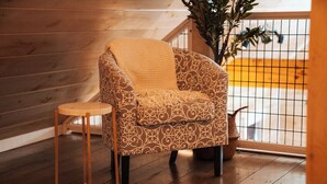 Un sillón estampado con una manta, un taburete de madera y una planta en maceta en una habitación con suelo de madera y barandilla.
