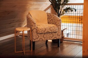 A patterned armchair with a throw blanket, a small wooden stool, and a potted plant in a room with wooden flooring and a railing.