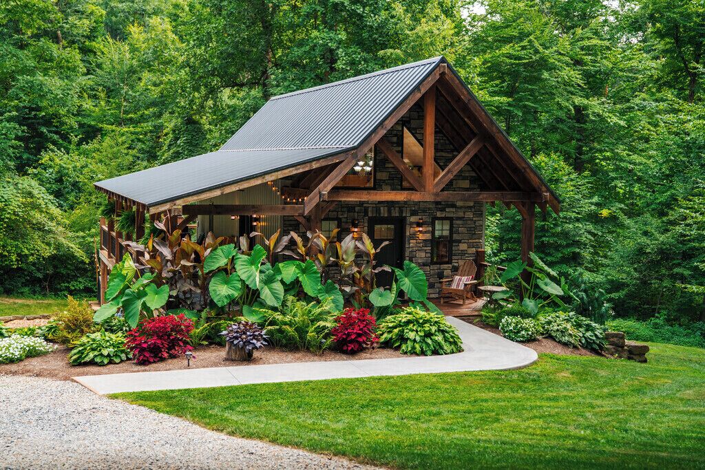 A rustic cabin with a sloped roof, surrounded by lush greenery and a variety of plants.