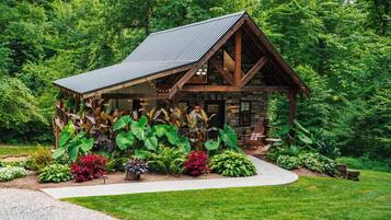A rustic cabin with a sloped roof, surrounded by lush greenery and a variety of plants.