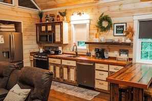 A wooden interior with a kitchen area, including a refrigerator, stove, and microwave. There is a wooden dining table and a couch with a cushion.