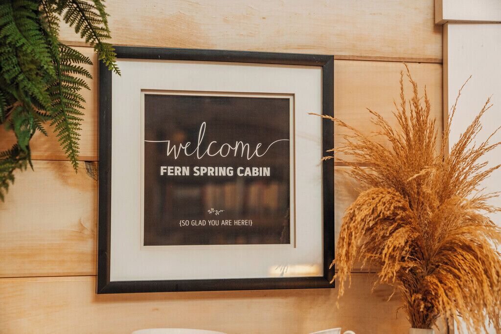 A framed welcome sign for Fern Spring Cabin with a simple message and a decorative plant beside it.