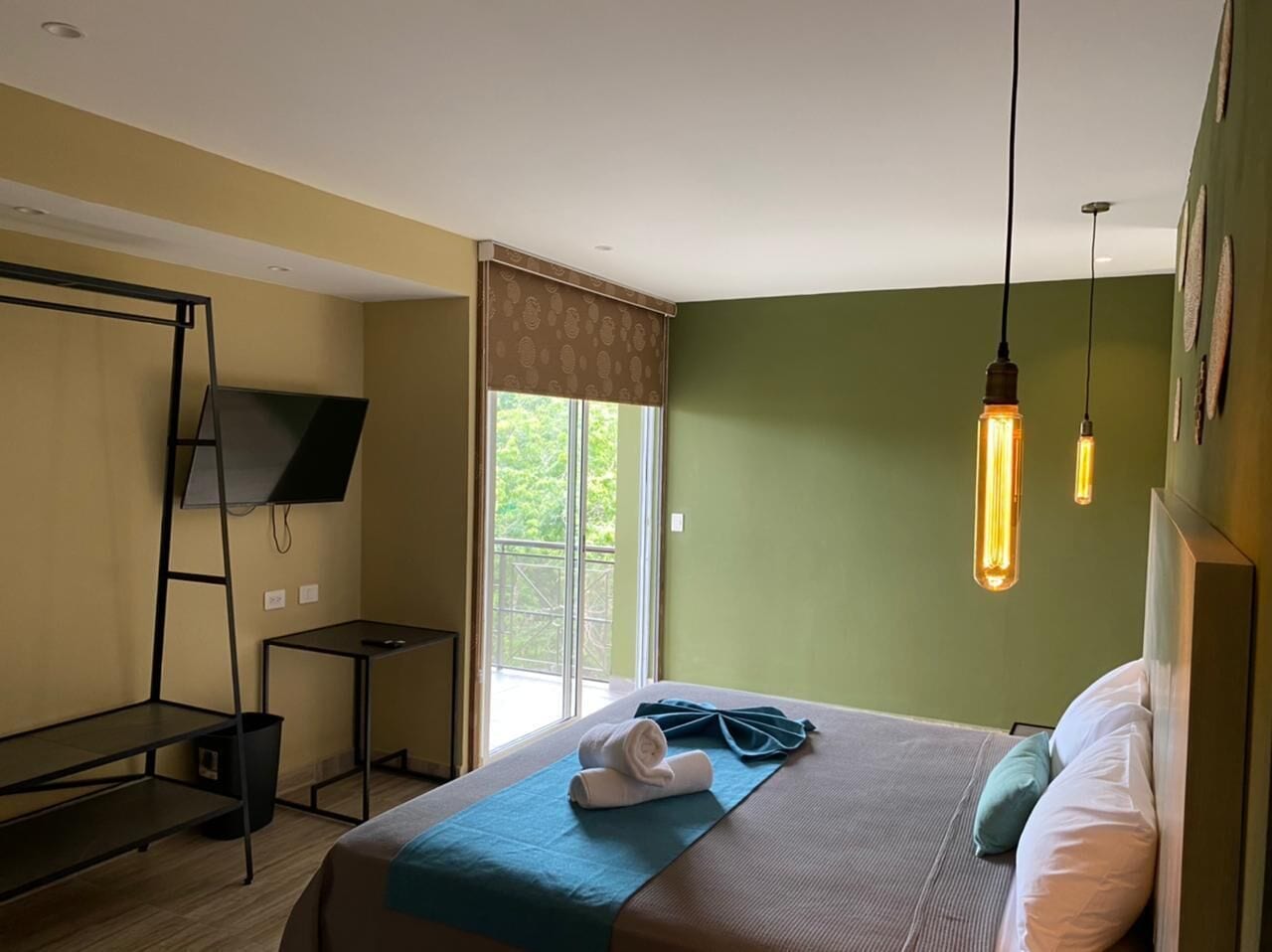 A hotel room with a bed, a TV on a stand, a window with curtains, and hanging light fixtures.