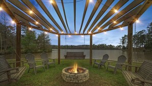 A wooden pergola with string lights, a fire pit, and Adirondack chairs around a lake.