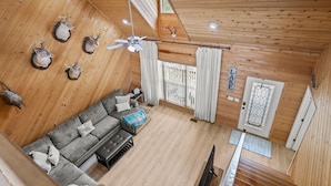 A wooden cabin interior with a sofa, ceiling fan, mounted deer heads, and a door with a geometric pattern.
