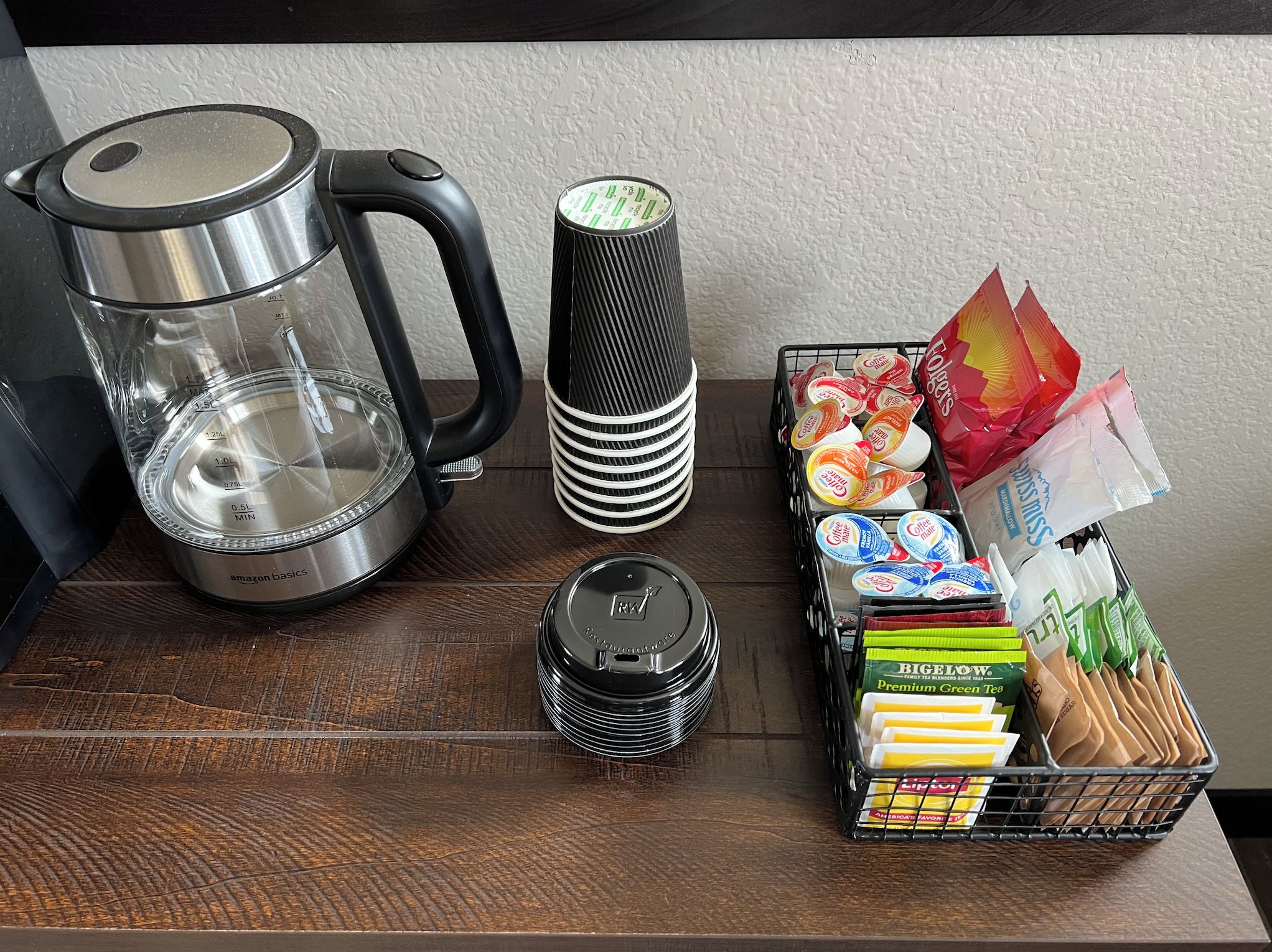 Un plan de travail de cuisine avec une machine à café, une bouilloire, une pile de couvercles noirs, une pile de couvercles blancs et un panier contenant des dosettes de café et des sachets de thé.