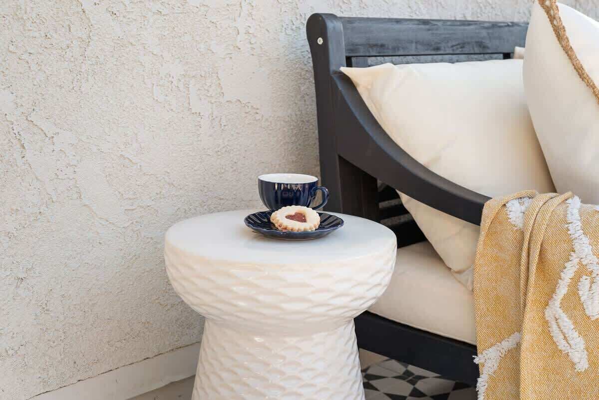 A white textured stool with a blue mug and a cookie on a small table beside a dark wooden chair with a beige cushion.