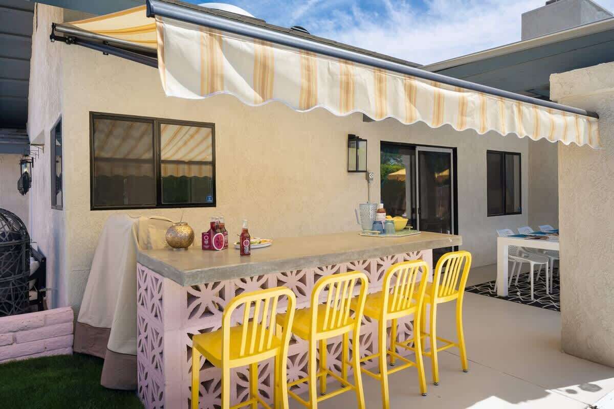 Une terrasse extérieure avec un coin bar, des chaises jaunes et un auvent à rayures.
