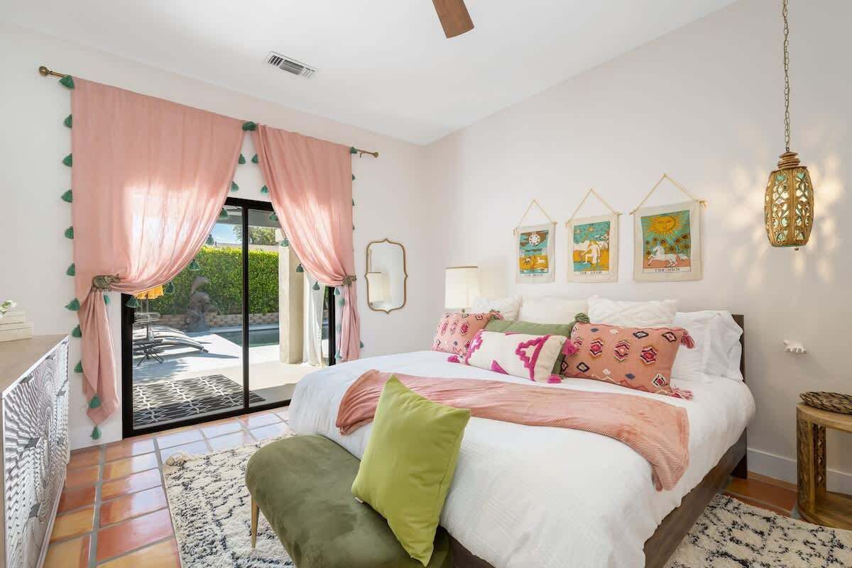 Un dormitorio con cama, almohadas, una mesita de noche, una lámpara, una ventana con cortinas y una pared decorada con cuadros.