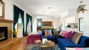 Ein modernes Wohnzimmer mit einem blauen Sofa, einem roten Hocker und einem gemusterten Teppich. Im Hintergrund befindet sich eine Küche mit weißen Schränken und einem Essbereich.