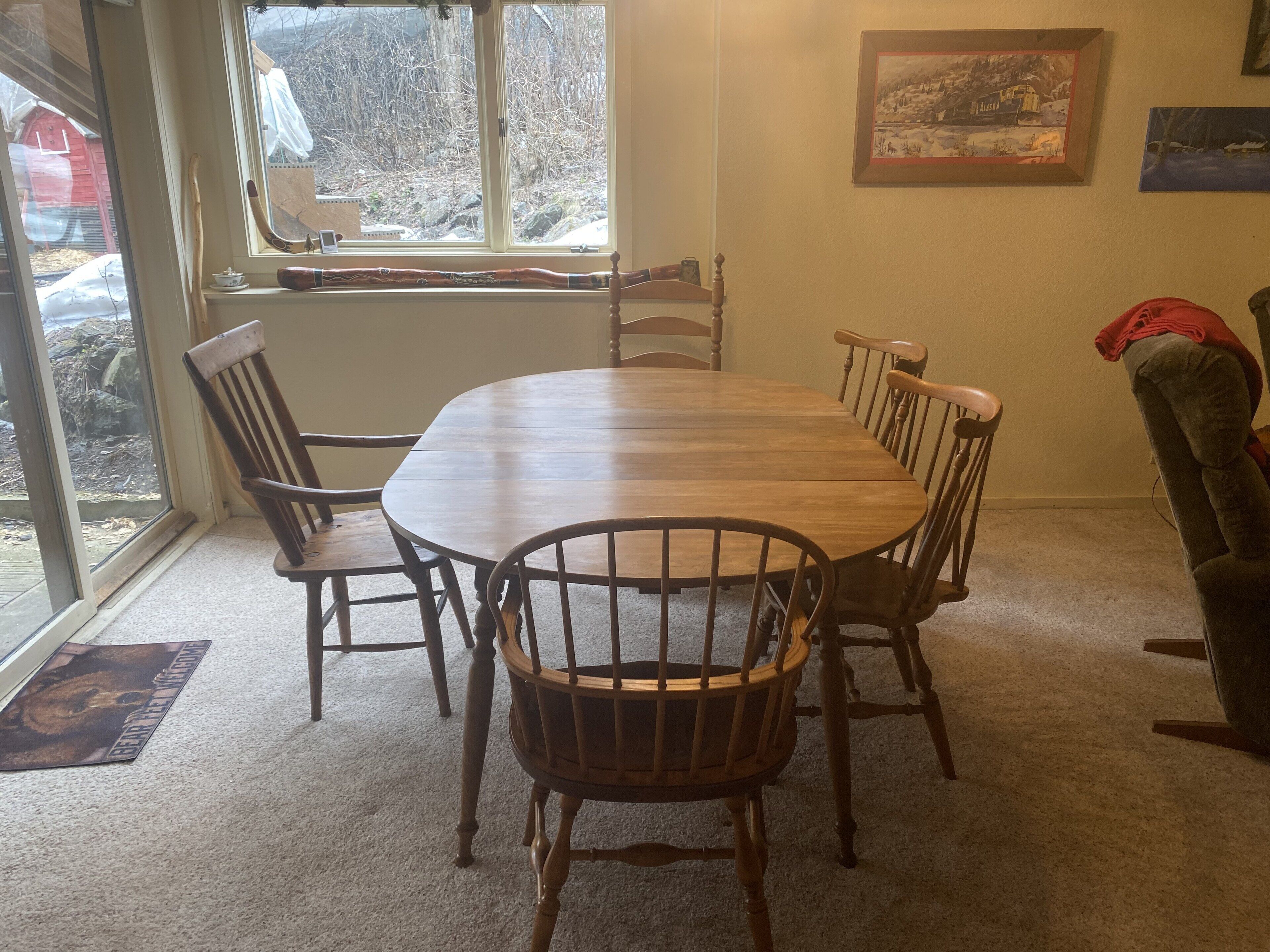 Un coin repas avec une table et des chaises en bois, une fenêtre donnant sur les arbres et un tableau encadré au mur.