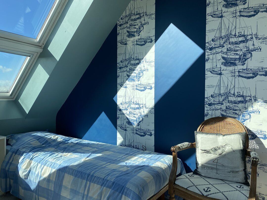 A bedroom with a skylight, a bed with a blue checkered bedspread, a chair with a cushion, and wallpaper with a nautical theme.